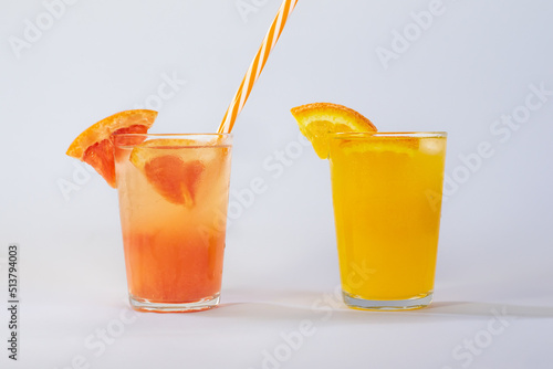 Photo of a glass of grapefruit juice with a red and white straw and a glass of orange juice on a white background