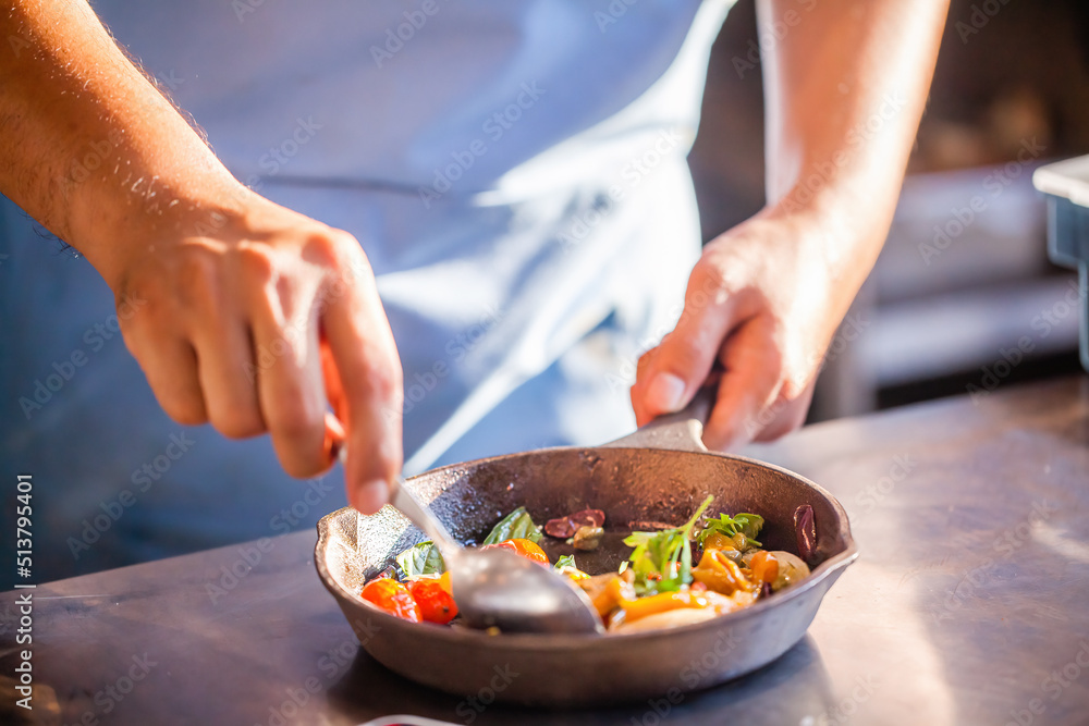 Chef cooking food in restaurant kitchen. Gourmet chef in uniform preparing meal, close up. Male in apron stirs with a spoon vegetables in frying pan on kitchen counter in luxury fine dining restaurant
