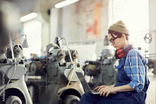 Concentrated young female production engineer in khaki beanie hat and blue overall sitting in workshop with lathes in background and analyzing sketch on laptop