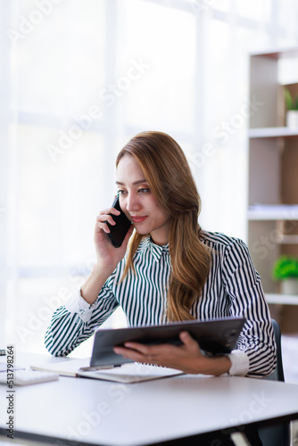 Beautiful young Asian woman work at home with laptop and talking on smartphone and smiling. Asian Female working on computer and using cell mobile phone while sitting at home