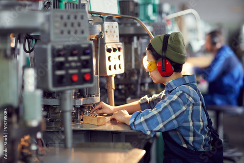 Busy tomboy in hat and ear protectors sitting at desk and processing small details on lathe at factory