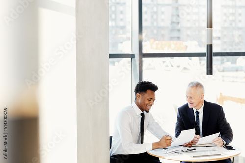 Smiling multi-ethnic business partners in formal outfits sitting at round table and viewing business papers together