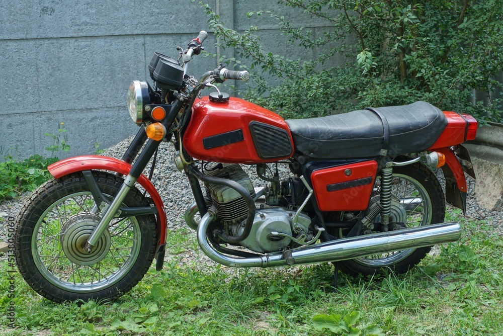 an old metal red color powerful popular motorcycle released in the soviet union stands in the summer on the street on green grass near a concrete fence