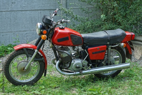 an old metal red color powerful popular motorcycle released in the soviet union stands in the summer on the street on green grass near a concrete fence