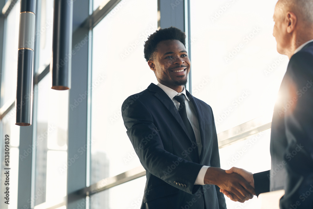 Fototapeta premium Cheerful handsome young Black businessman in suit standing at panoramic window and making handshake with business partner in lobby of company