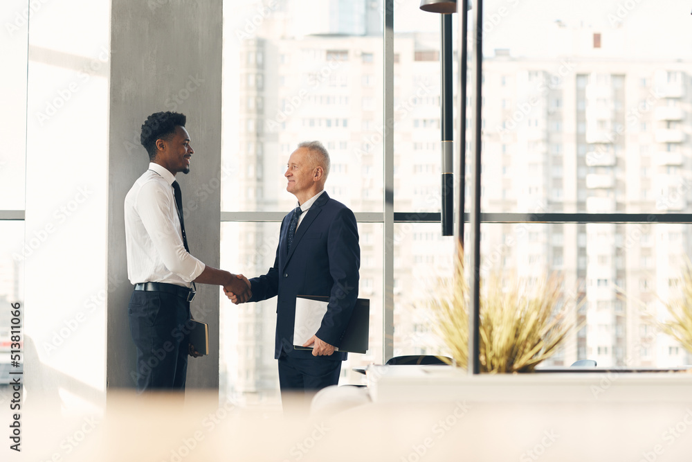 Senior recruiter with papers shaking hand of young Afro-American specialist while hiring him for work Senior recruiter with papers shaking hand of young Afro-American specialist while hiring him for work
