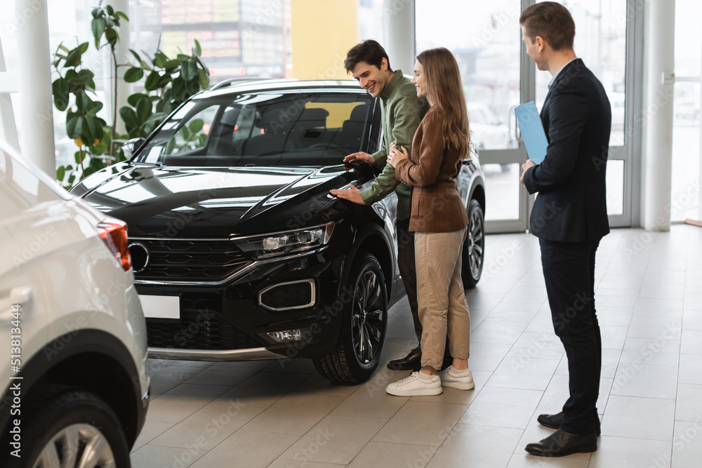 Cheery millennial couple speaking to car salesman about buying or renting new vehicle at dealership, copy space