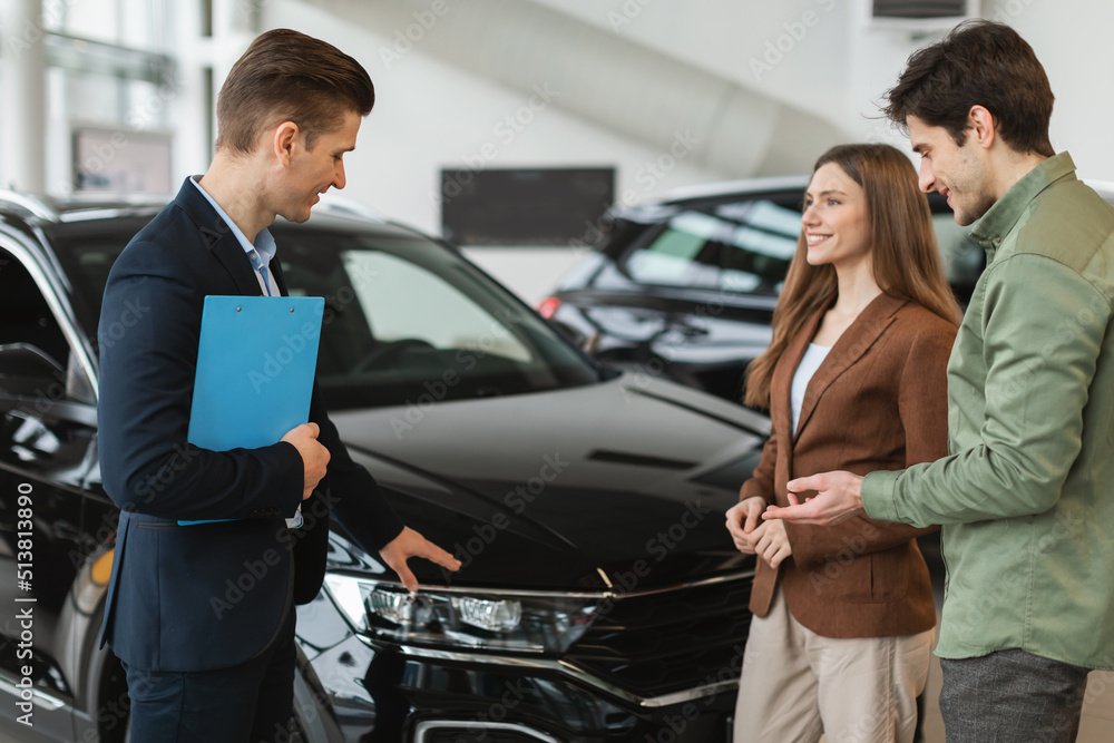 Positive young spouses speaking to salesman about purchasing new auto at dealership shop. Car purchase or rental Positive young spouses speaking to salesman about purchasing new auto at dealership shop. Car purchase or rental