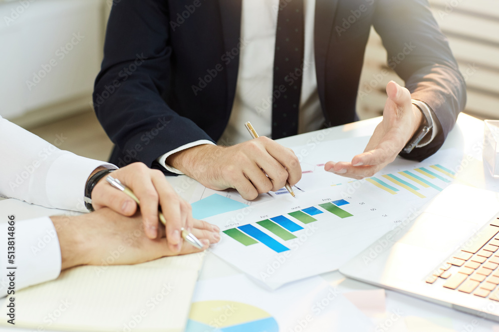 Close-up of unrecognizable businessman in black suit sitting at desk with analytical papers and discussing financial issues with business partner Close-up of unrecognizable businessman in black suit sitting at desk with analytical papers and discussing financial issues with business partner