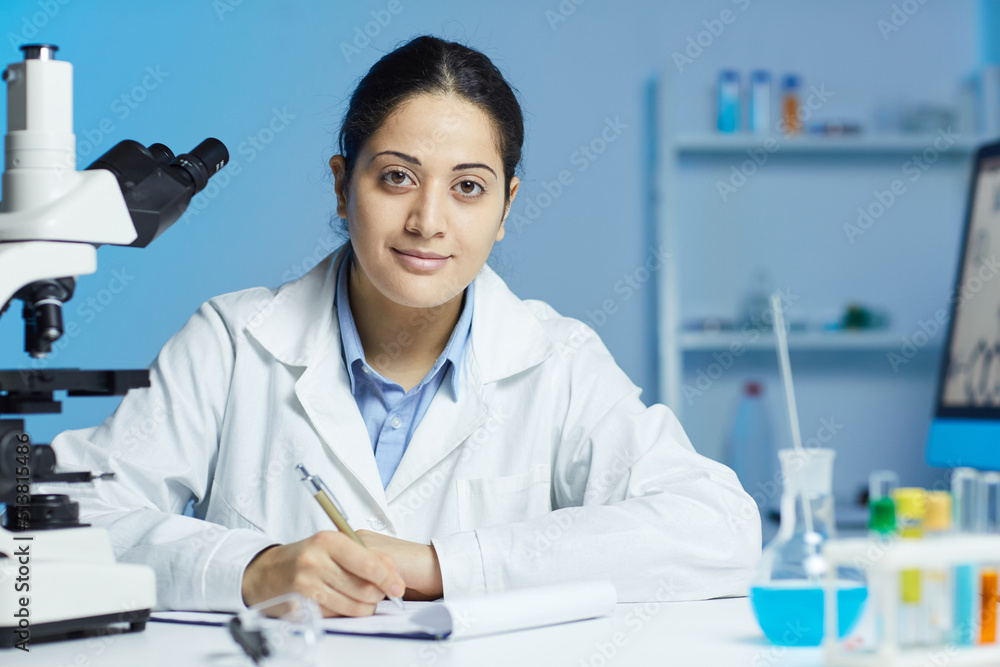 Portrait of content Indian female intern in white coat sitting at desk and writing down information on experiment in laboratory Portrait of content Indian female intern in white coat sitting at desk and writing down information on experiment in laboratory