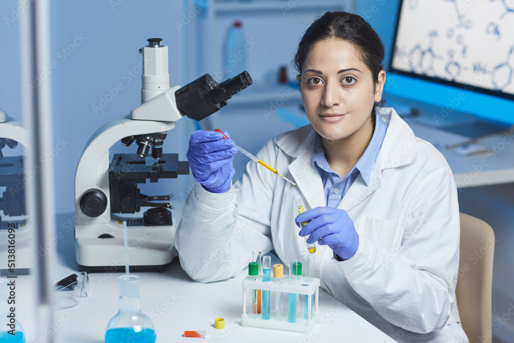 Portrait of content young Indian female pharmacist in lab coat and surgical gloves sitting at desk and mixing reagents in test tube using pipette Portrait of content young Indian female pharmacist in lab coat and surgical gloves sitting at desk and mixing reagents in test tube using pipette