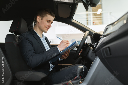Positive young auto salesman sitting inside new car, making checkup, taking notes during test drive at dealership © Prostock-studio