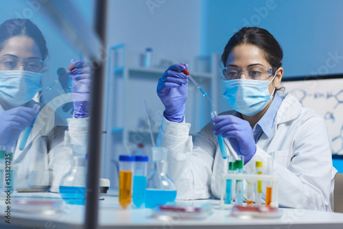 Concentrated young Indian female laboratory worker in mask and safety goggles sitting at desk and adding blue liquid into test tube