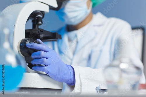 Close-up of unrecognizable biologist in rubber gloves and white coat sitting at desk and adjusting microscope in laboratory