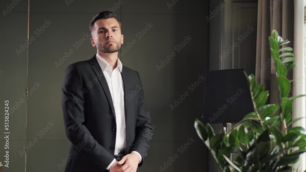 Stylish bearded man stands in front of a window and going to a business meeting. Close up view of man adjusts his suit. Businessman put suit on during morning routine at home, preparing to go to work. Stylish bearded man stands in front of a window and going to a business meeting. Close up view of man adjusts his suit. Businessman put suit on during morning routine at home, preparing to go to work.