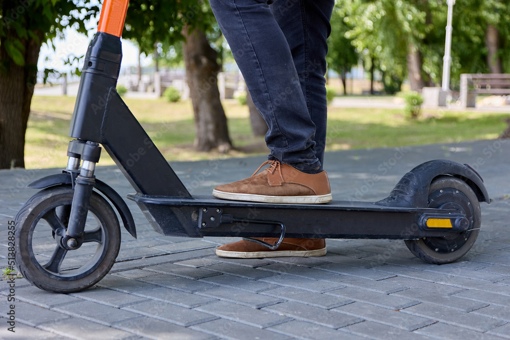 Obraz premium Close up of men's legs ready to start in a scooter race. Against the background of trees in a city park, a photo of a modern electric scooter and a man riding on paving slabs