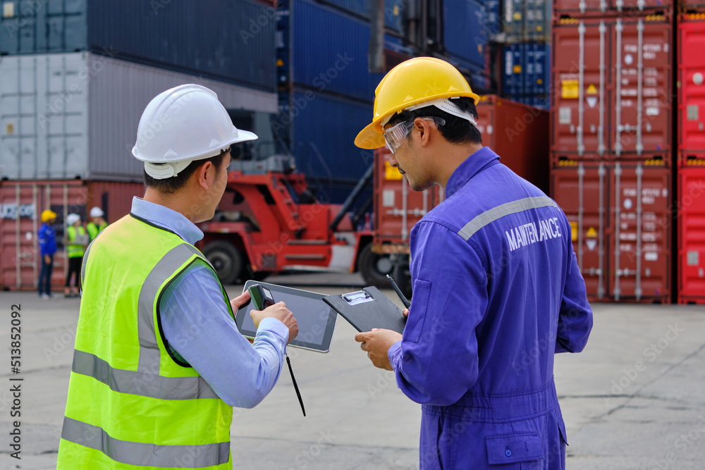 Two professional Asian male workers in safety uniforms and hard hats work at a logistics terminal with many stacks of containers, loading control shipping goods for the cargo transportation industry.