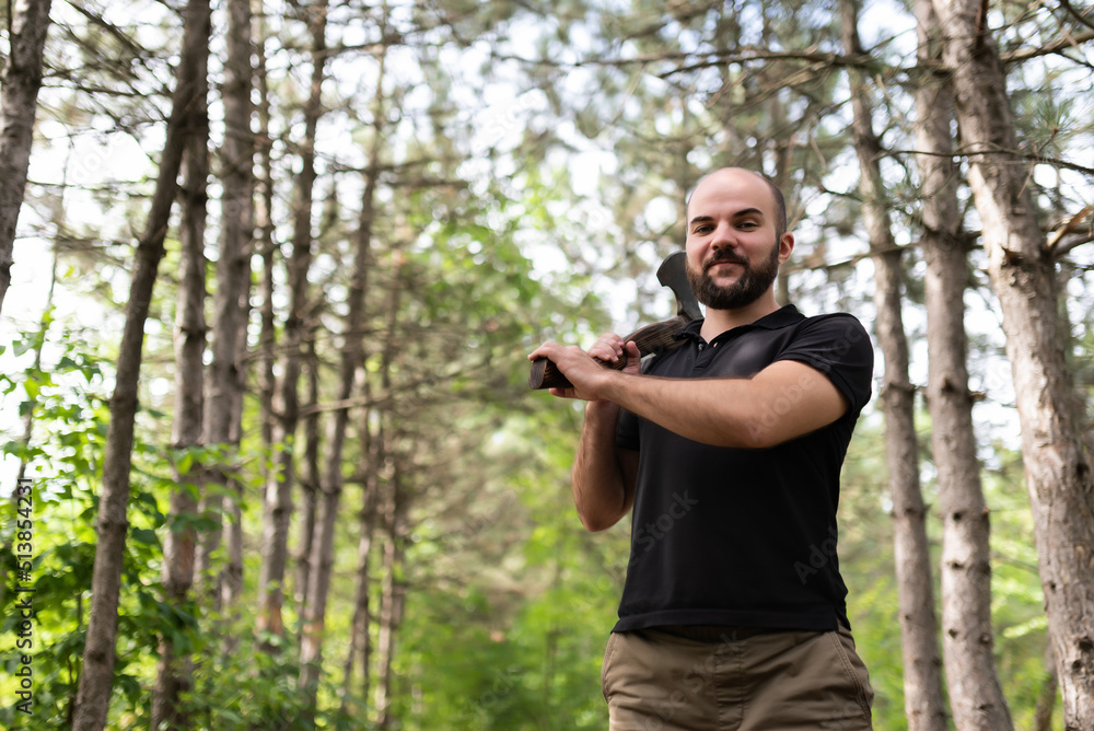 Bearded guy in a black T-shirt stands in a summer forest with an ax in his hands. Looks into the camera. Lumberjack posing for the camera. Chopping wood. Natural landscape Bearded guy in a black T-shirt stands in a summer forest with an ax in his hands. Looks into the camera. Lumberjack posing for the camera. Chopping wood. Natural landscape