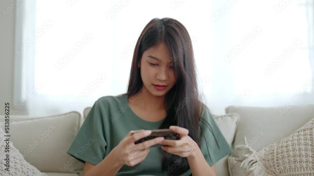 Happy young asian woman relax on comfortable couch at home, playing online gaming on smartphone, smiling girl use cellphone chatting, browse wireless internet on gadget, shopping online from home Happy young asian woman relax on comfortable couch at home, playing online gaming on smartphone, smiling girl use cellphone chatting, browse wireless internet on gadget, shopping online from home