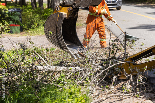 Closeup view on hydraulic arm of an excavator at work clearing tree branches and debris from the highway after a storm, man with rake in background.