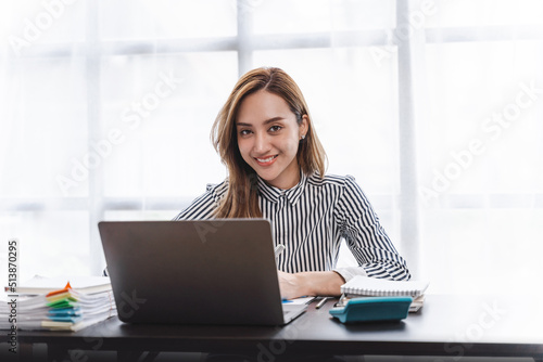 Looking to camera, Young confident Asian businesswoman working at office desk and typing with a laptop, office shelves on background.