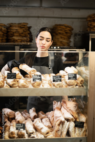 Beautiful young woman working in a bakery shop, bakery owner at work, showcase with pastry assortment with price tags, small business