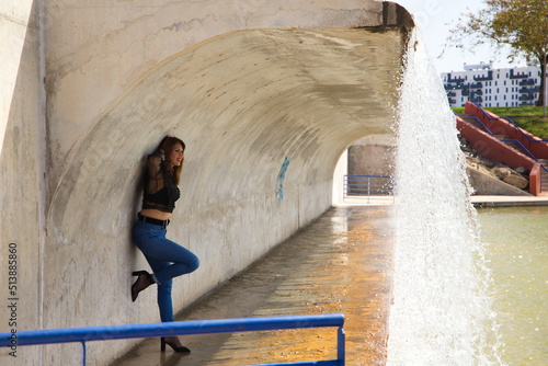 Beautiful, fashionably dressed mature woman under a large waterfall in the park. The woman is posing with her arms outstretched on the wall in a sensual way. Fashion and beauty concept.