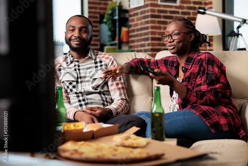 African american people switching tv channels and eating takeaway meal from fast food delivery at home. Watching movie on television program and enjoying takeout food with beer bottles.