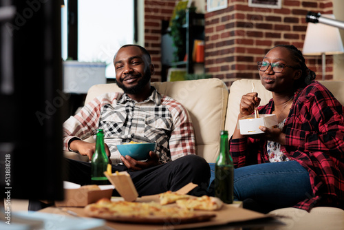 African american couple eating takeaway meal with noodles and chips to watch movie on television. Leisure activity together with film on tv and fast food delivery package, having fun.