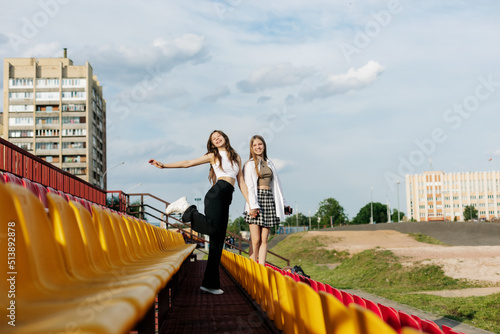 Two teenage girls walk together through the stands of the school stadium, talking, holding hands, best friends return home after training © Sviatlana