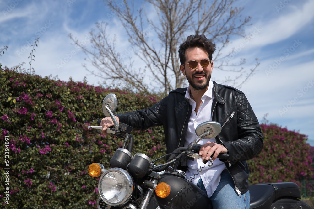 Handsome young man with beard, sunglasses, leather jacket, white shirt and jeans, riding a large displacement motorcycle. Concept beauty, fashion, trend, motor, motorcycle, naked.
