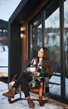 Young woman resting on terrace of modern barn house in the mountains. Happy female tourist sitting in chair, holding cup of tea, enjoying in new cottage in winter.