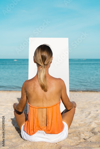 A blonde woman sits on a sandy beach in front of a white canvas.  The sea and blue sky is in the background.