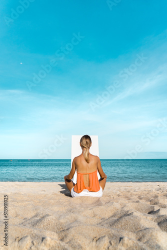 A blonde woman sits on a sandy beach in front of a white canvas.  The sea and blue sky is in the background.