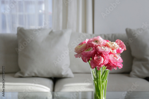 Close up shot of glass coffee table with bouquet of beautiful bicolor ranunculus flowers in a vase on foreground and gray textile couch on the background. Copy space, close up, natural light.