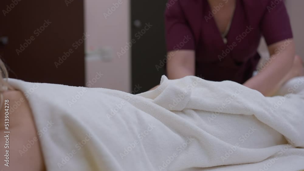 Unrecognizable woman covered with white towel lying in spa salon with blurred masseur massaging massaging legs at background. Relaxed carefree client enjoying massage in beauty center Unrecognizable woman covered with white towel lying in spa salon with blurred masseur massaging massaging legs at background. Relaxed carefree client enjoying massage in beauty center