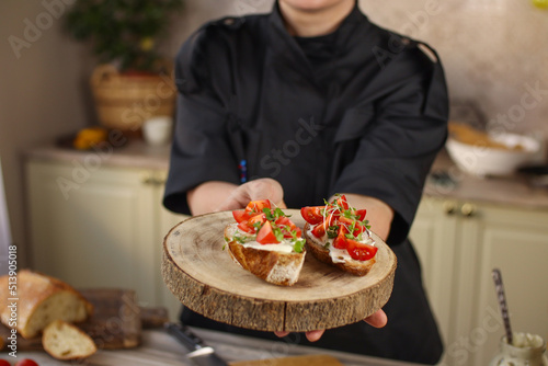 Female chef preparing breakfast in a bright country kitchen. He cuts the bread and spreads it with cheese. Kitchen table with flowers and kitchen board, bread, tomatoes, greens