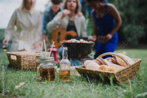 Close-up of picnic near lake in summer, young friends in background eating and playing guitar.