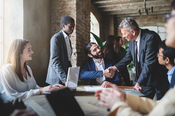 Two businessmen make a deal by shaking hands sitting at a table together with other multiracial employees - business lifestyle and agreements concept