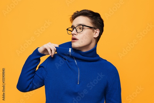 an attractive young man stands on an orange background in a blue zip-up jacket and black eyeglasses and pulls aside the neck of the jacket