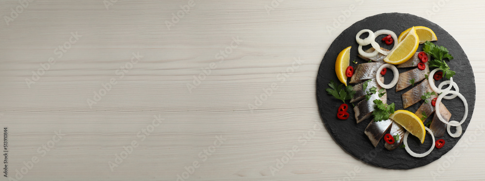 Slate plate with sliced salted herring fillet, parsley, chili pepper, onion rings and lemon on light beige wooden table, top view with space for text. Banner design