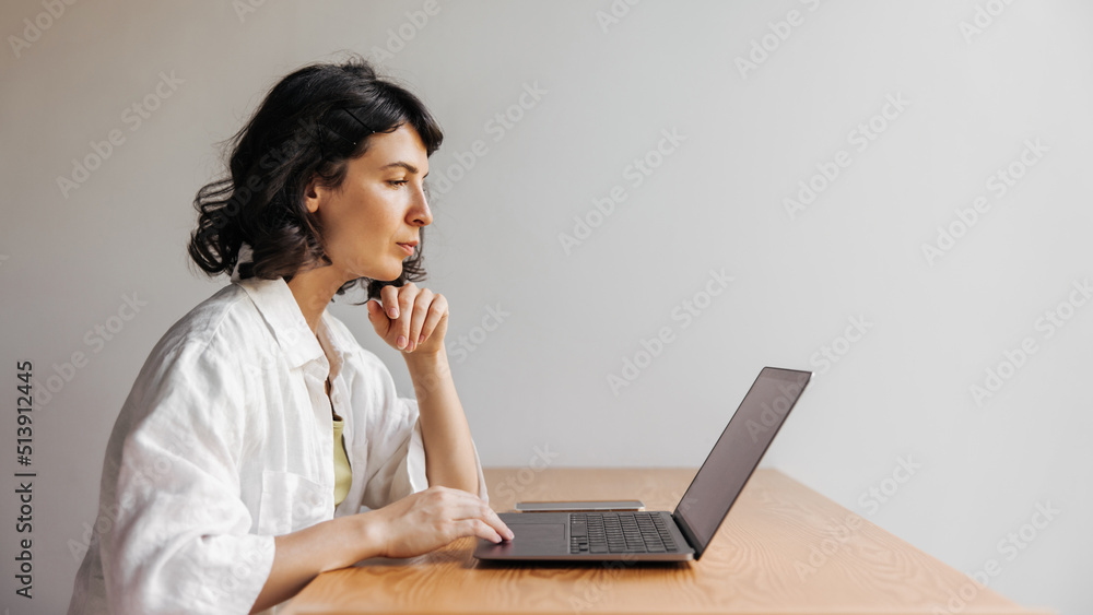 Focused young caucasian woman is using modern smartphone sitting at table. Brunette in white shirt makes online purchases. Technology concept Focused young caucasian woman is using modern smartphone sitting at table. Brunette in white shirt makes online purchases. Technology concept