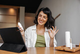 Attractive young caucasian woman smiling looking into tablet screen sitting at table. Brunette wearing casual clothes shows ok sign. Technology concept