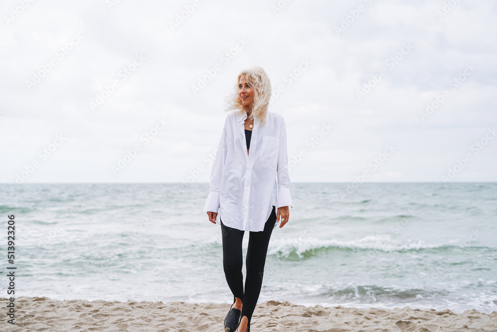 Portrait of Elegant blonde woman in white shirt on sand beach at storm sea at windy weater Portrait of Elegant blonde woman in white shirt on sand beach at storm sea at windy weater