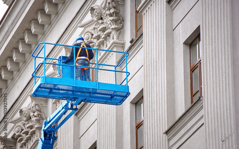Construction worker in lift bucket of crane restore and repair historic facade of building. Man in cradle, restoring plaster decoration on facade. Workers painting building, renovating and repair work Construction worker in lift bucket of crane restore and repair historic facade of building. Man in cradle, restoring plaster decoration on facade. Workers painting building, renovating and repair work
