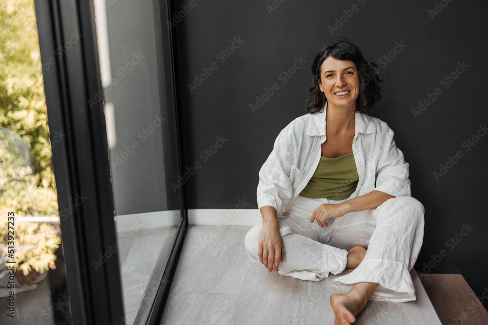 Happy young caucasian woman sits on floor by window alone and looks at camera. Brunette spends her leisure time at home during day. Relaxation concept Happy young caucasian woman sits on floor by window alone and looks at camera. Brunette spends her leisure time at home during day. Relaxation concept