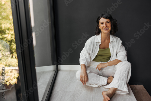 Happy young caucasian woman sits on floor by window alone and looks at camera. Brunette spends her leisure time at home during day. Relaxation concept