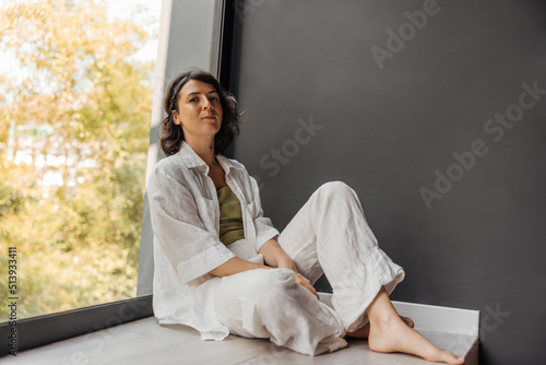 Pleasant young caucasian brunette woman sits on floor leaning against window in room. Girl looks at camera, wears casual clothes. Leisure lifestyle concept