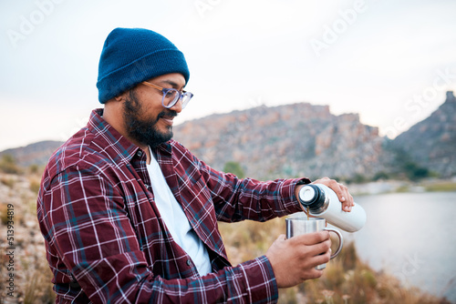 An adult man pours coffee on hiking trip in the mountains with lake view