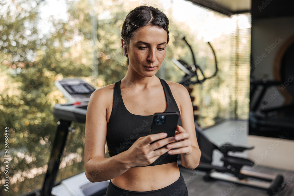 Calm young caucasian girl uses smartphone in gym. Brunette wears black sports top. Healthy lifestyle, technology concept